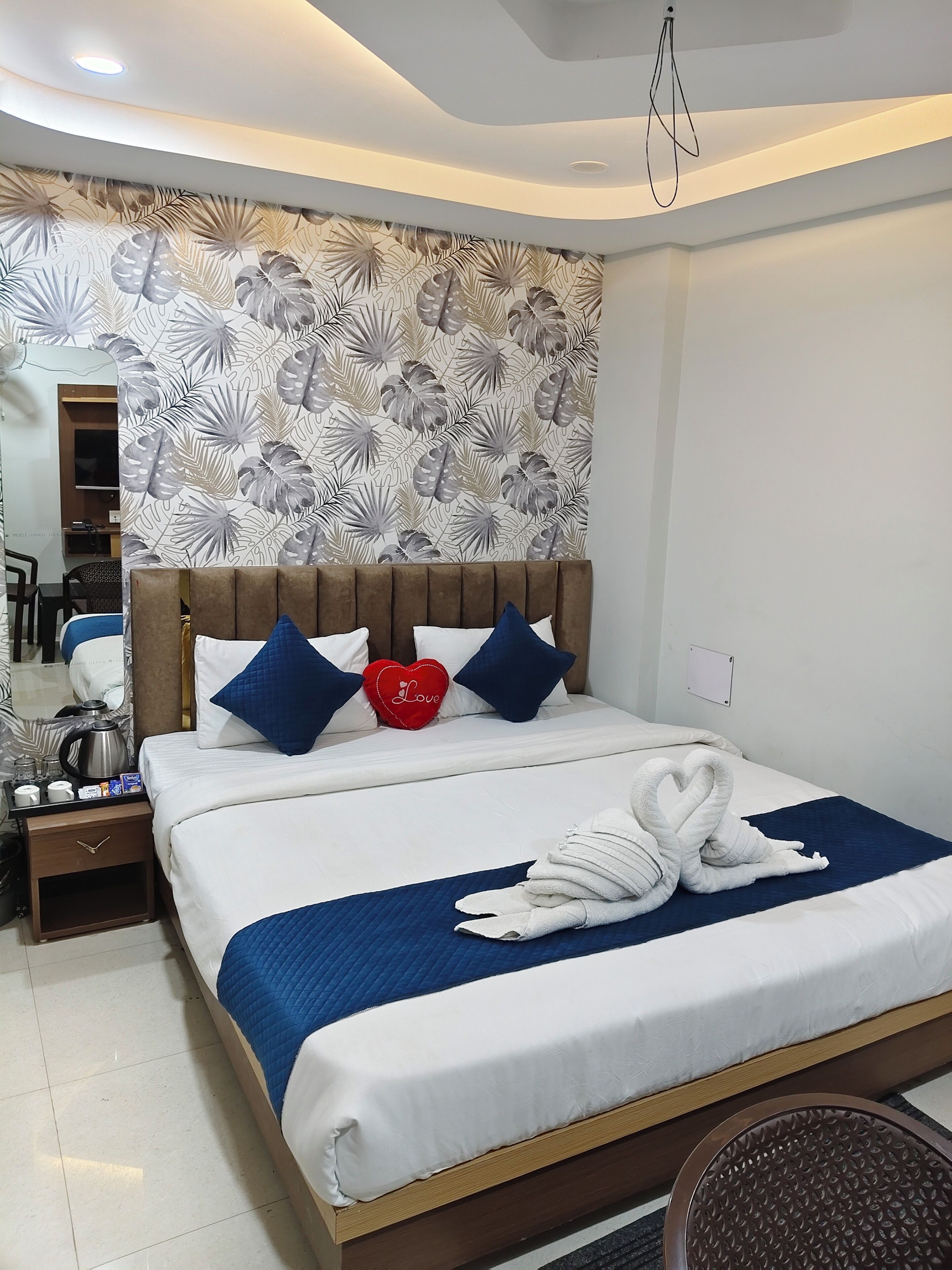 Habitación de hotel con cama, mesita de noche, espejo, pared con diseño de hojas y luz en el techo.