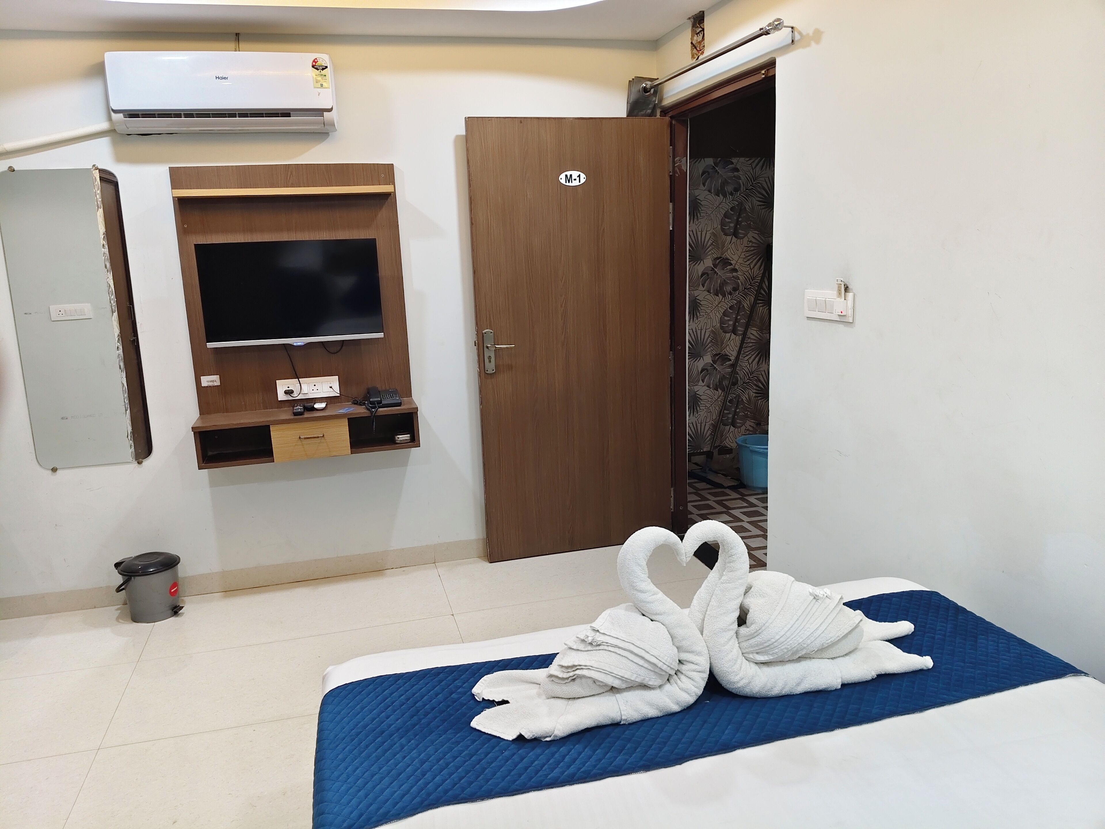 A hotel room with a bed, a TV mounted on the wall, an air conditioner, a door with a sign, and a trash bin.