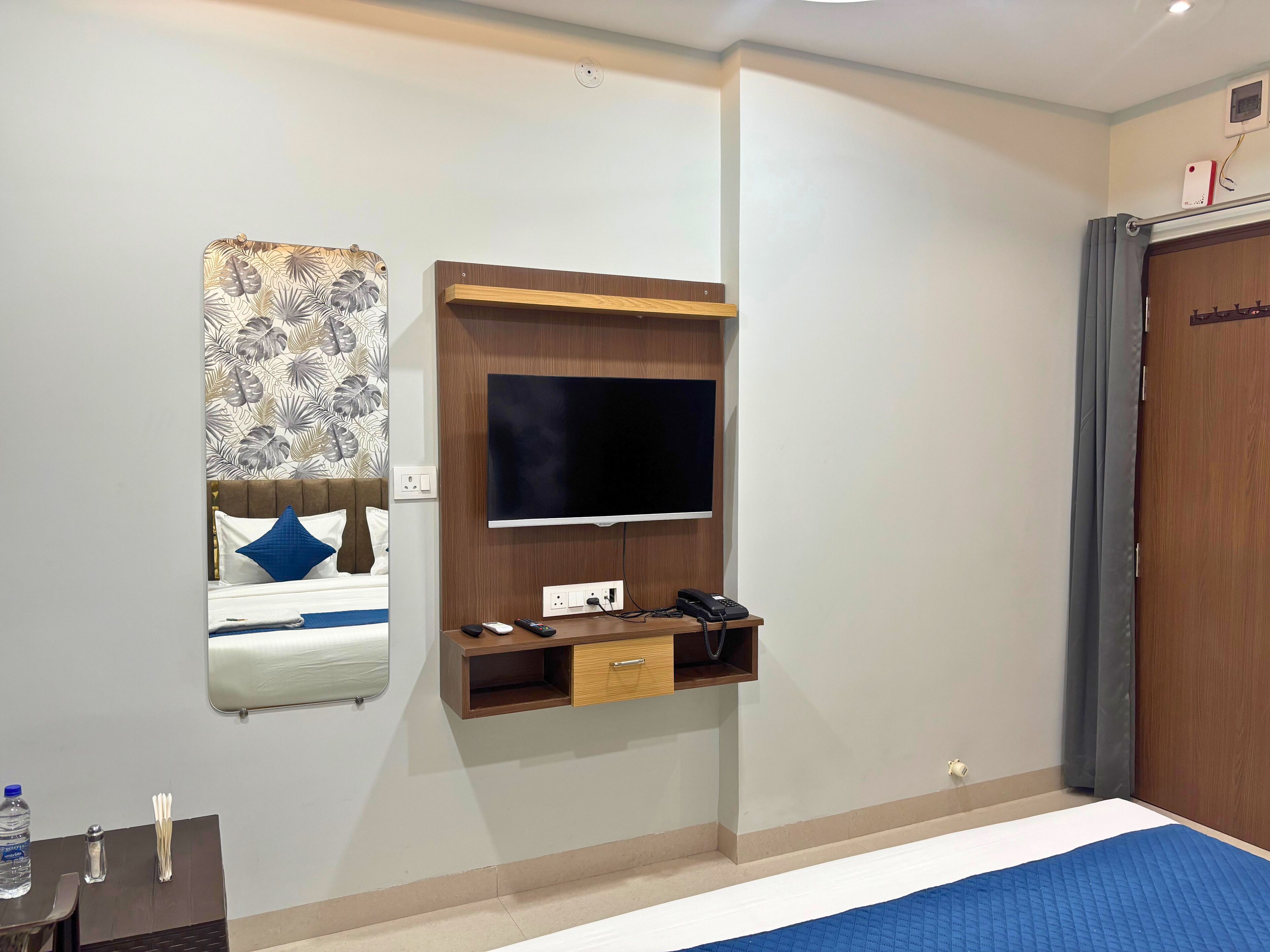 A hotel room with a bed, a TV mounted on the wall, a bedside table with a phone and remote controls, and a mirror with a floral pattern.