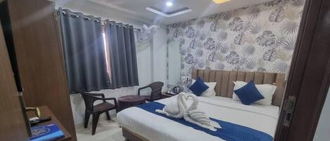 A hotel room with a bed, a desk, a chair, a fan, and a window with curtains.
