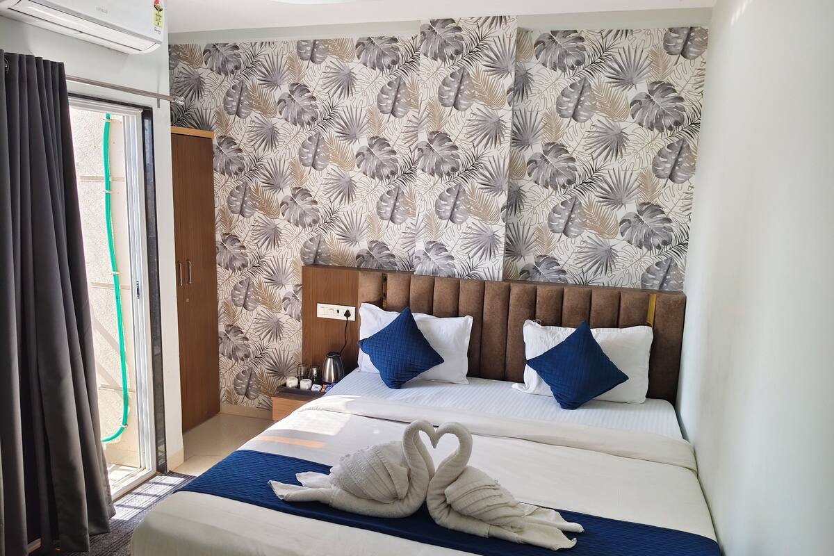 A hotel room with a bed, two pillows, a nightstand, a wall with a leaf pattern, and a window with curtains.