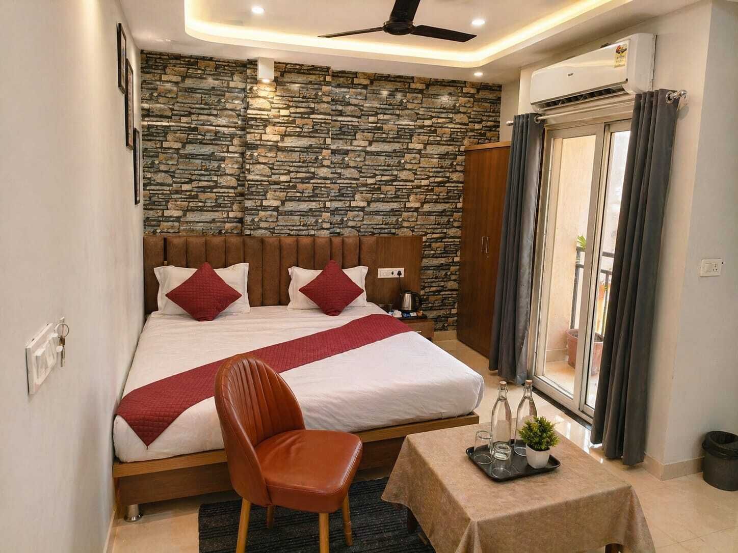 A bedroom with a stone wall, a bed with red pillows, a wooden headboard, a chair, a small table with a tray, and a window with curtains.