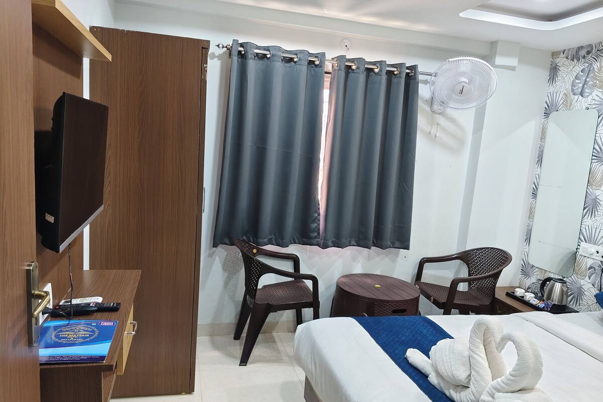 A hotel room with a bed, two chairs, a small table, a TV, a fan, and a curtain.