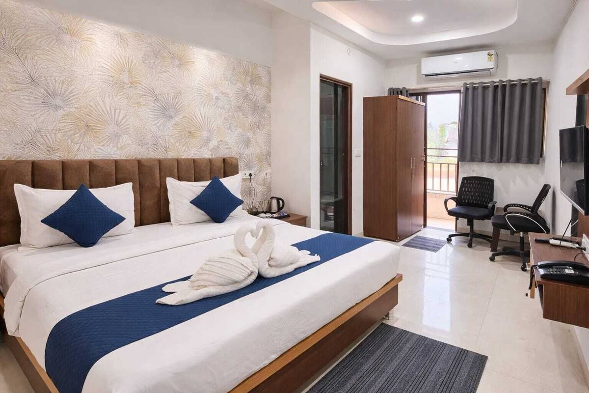 A hotel room with a large bed, a desk with a chair, a wardrobe, and an air conditioning unit.