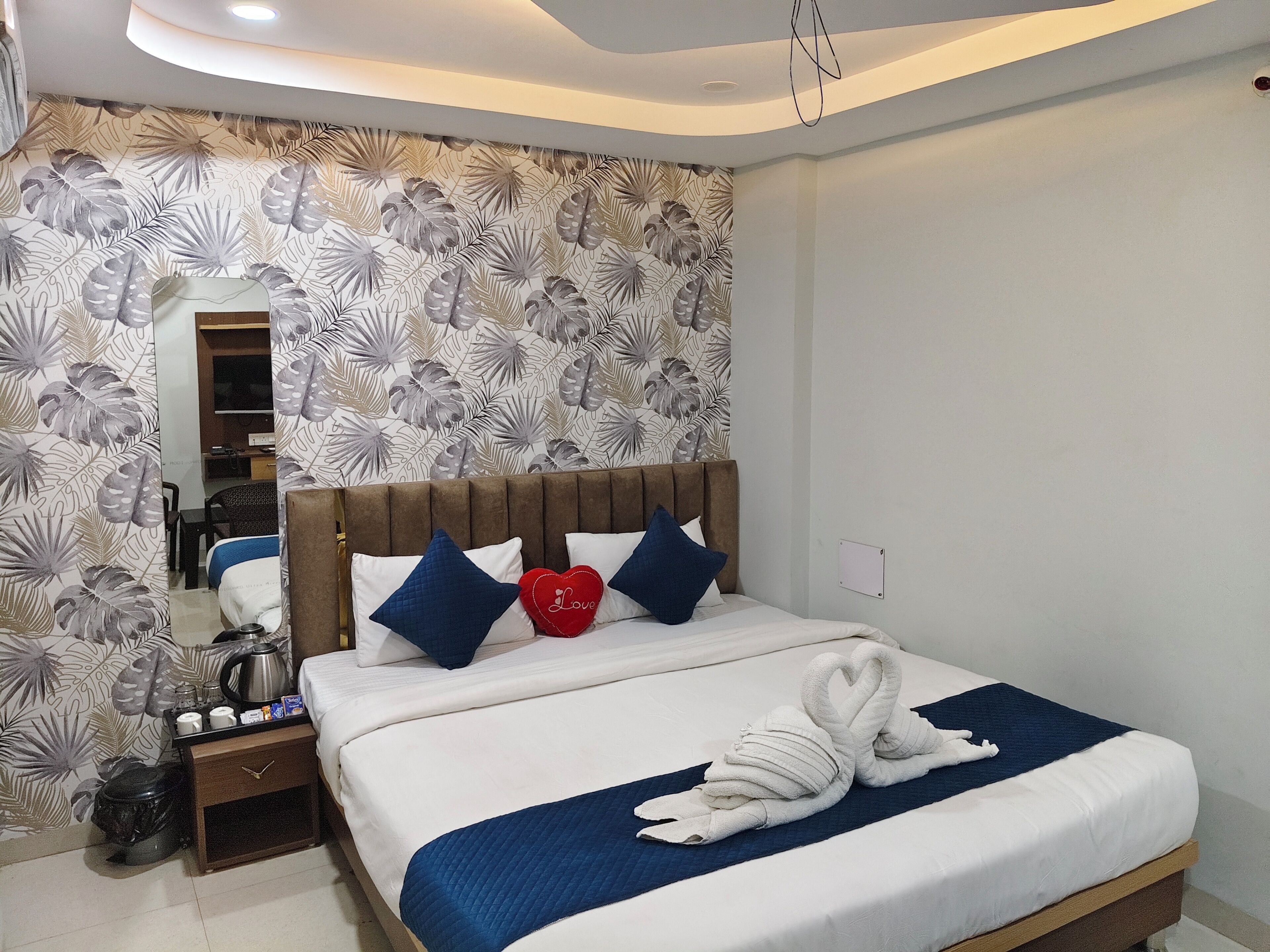 A neatly made bed with a blue blanket and a heart-shaped pillow, a wooden nightstand with a kettle, and a wall with a tropical leaf pattern.