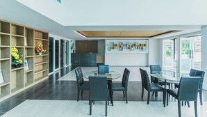 Ein modernes Interieur mit einem runden Glastisch, umgeben von Stühlen, einem Holzregal mit Blumen und einer Küche im Hintergrund.