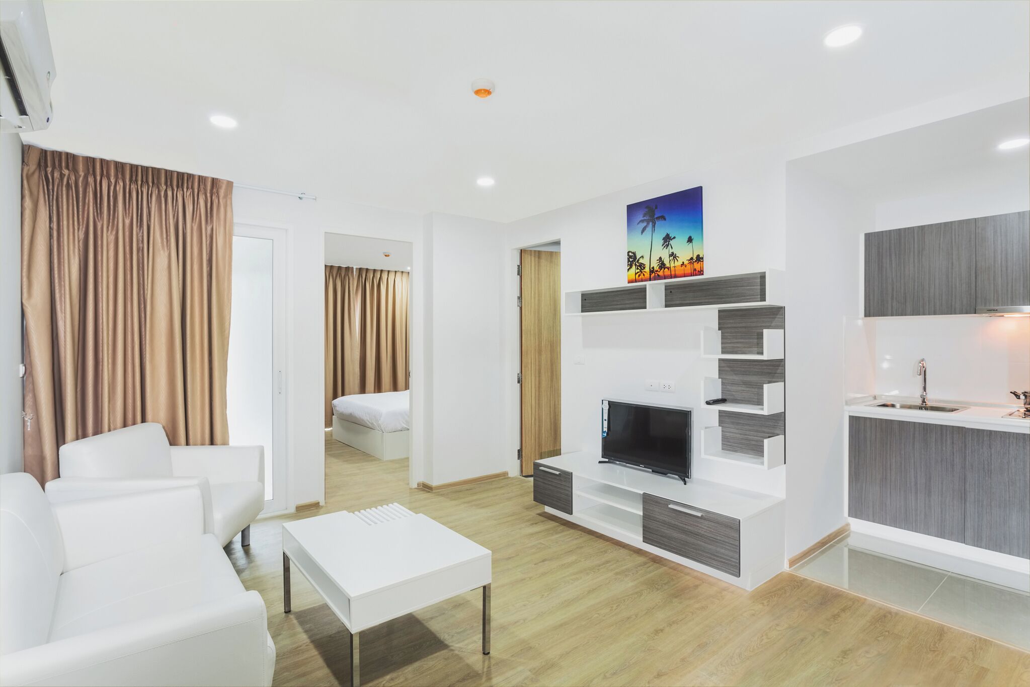 A modern living room with a flat-screen TV, white sofas, a coffee table, and a kitchen area with a sink and cabinets.