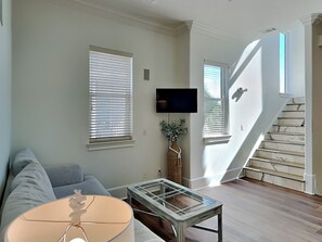 A living room with a sofa, a glass-top coffee table, a TV mounted on the wall, and a staircase leading to an upper floor.