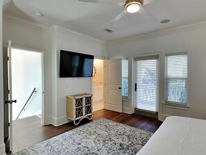 A bedroom with a ceiling fan, a TV mounted on the wall, a bed, a small cabinet, and a window with blinds.