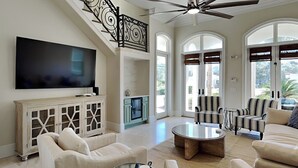 A spacious living room with a large flat-screen TV, comfortable seating, a ceiling fan, and large windows. - Blue Mountain Bella (Santa Rosa Beach)