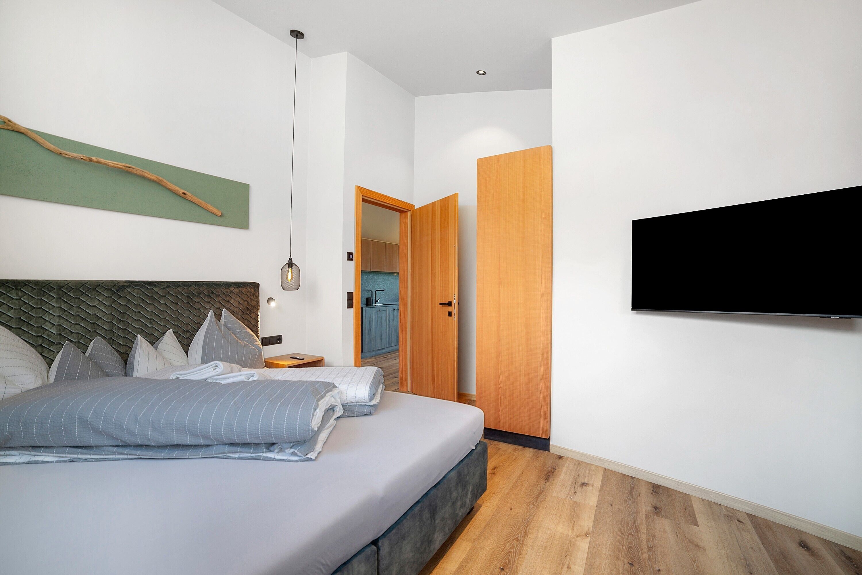 A modern bedroom with a bed, a TV, a wooden door, and a view into a kitchen.