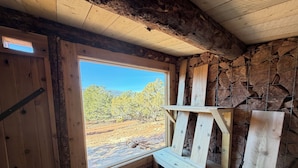A rustic cabin interior with a large window offering a view of a dry, rocky landscape with sparse trees.