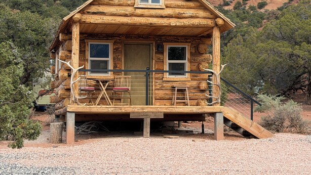 A rustic log cabin with a porch, two windows, and a small staircase leading up to the entrance.