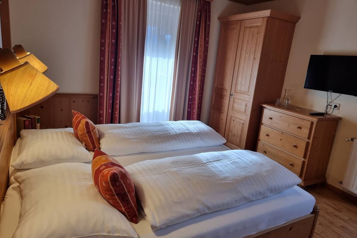A double bed with white linens, a wooden wardrobe, a desk with a lamp, and a television.