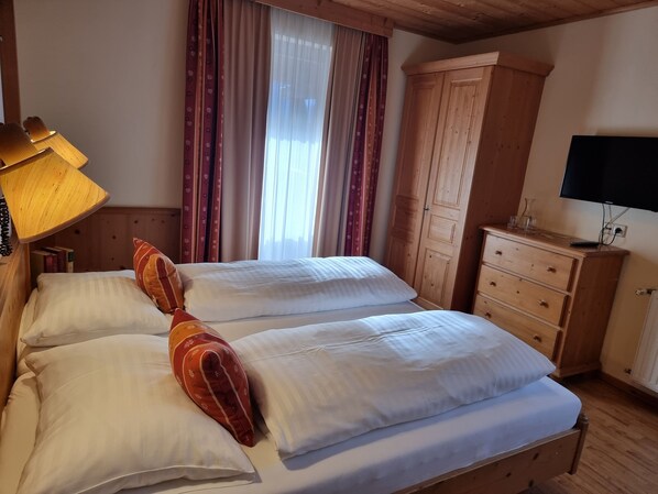 Un lit double avec du linge de lit blanc, une armoire en bois, un bureau avec une lampe et un téléviseur.