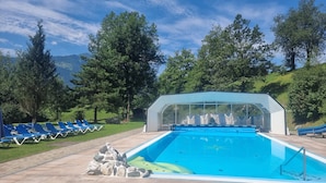 Una piscina con sillones de descanso y una zona de estar cubierta, rodeada de árboles y un paisaje montañoso.