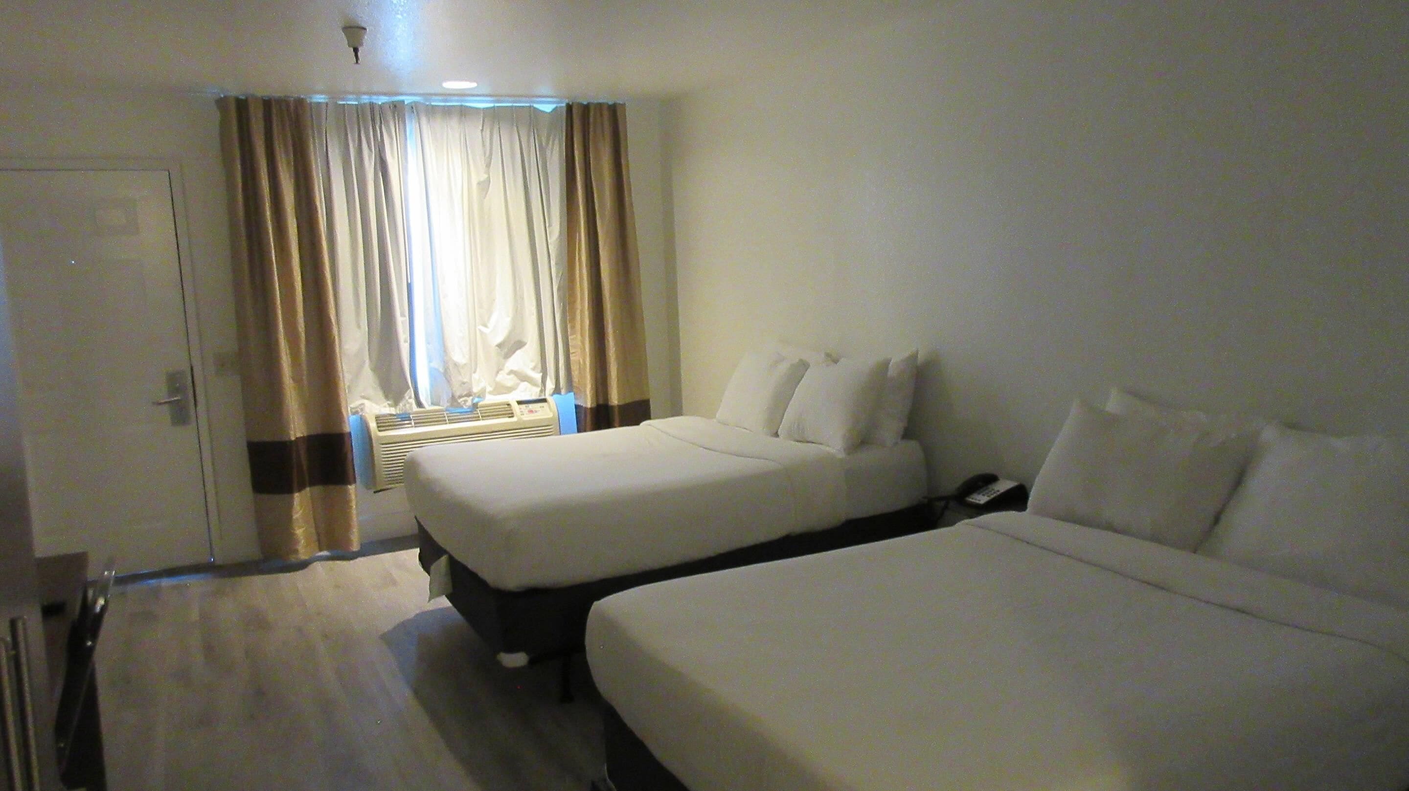 A hotel room with two beds, a bedside table, a lamp, and a window with curtains.