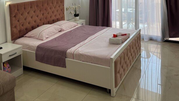A modern bedroom with a large bed, a bedside table, a nightstand, and a view of the outside through sheer curtains. - Cityheart Apartments Ulcinj, Montenegro (Ulcinj Municipality)