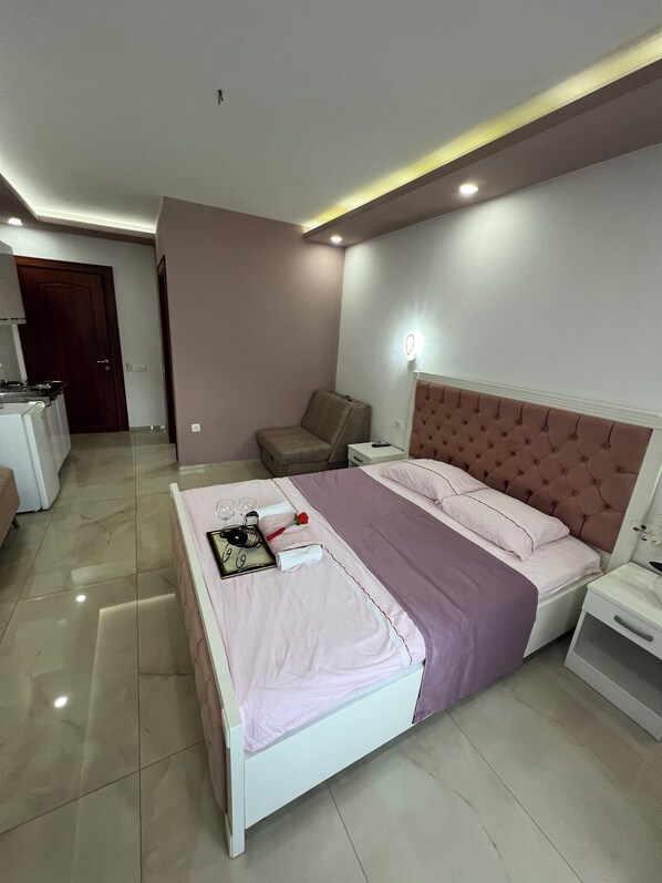 A modern bedroom with a bed, bedside tables, a nightstand, a chair, and a kitchenette area.