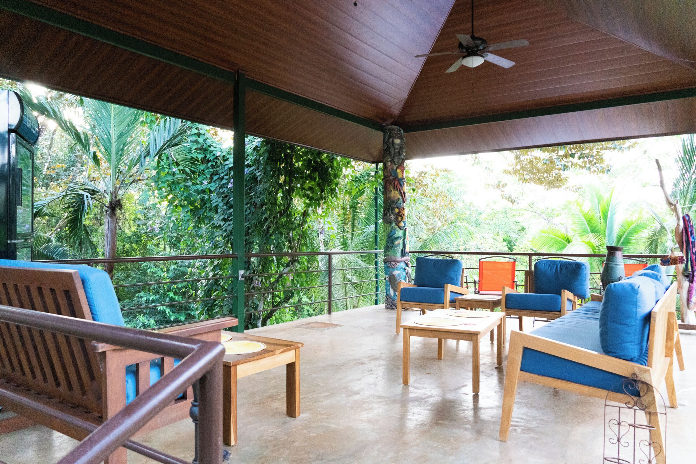 A covered outdoor seating area with wooden furniture and a view of lush greenery.