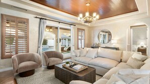 Ein geräumiges Wohnzimmer mit einem beigefarbenen Sofa, zwei Sesseln, einem Couchtisch und einer Deckenleuchte. Große Fenster mit Holzjalousien und Blick auf einen Außenpool sind zu sehen.