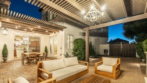 Ein möblierter Außenbereich mit Holzsofa, Sesseln und Couchtisch. Über dem Patio befindet sich eine Pergola mit Lichterketten, und es gibt einen Blick in den Garten.