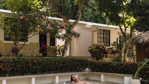 A pool area with a person in a bikini, surrounded by lush greenery and a building with a thatched roof. - Mística Natural  (Playa Conchal)