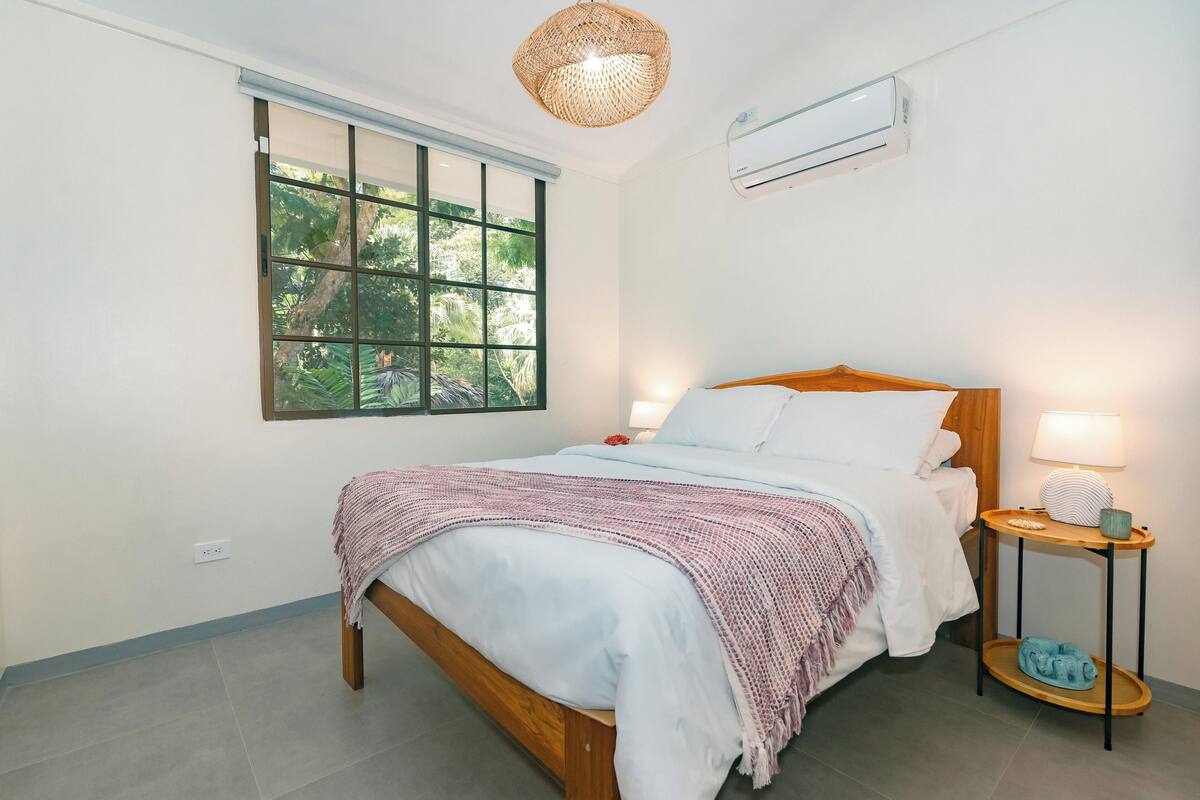 A neatly arranged bedroom with a wooden bed, a bedside table with a lamp, and a window with a view of greenery.