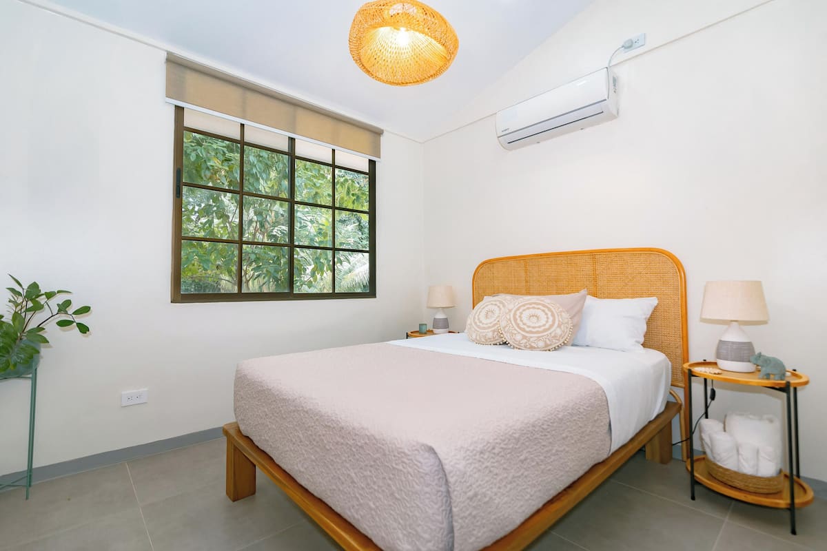 A bedroom with a bed, bedside tables, a window, an air conditioner, and a hanging light fixture.
