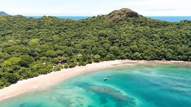An aerial view of a beach with clear turquoise waters, a sandy shore, and dense green forested hills in the background. - Mística Natural  (Playa Conchal)