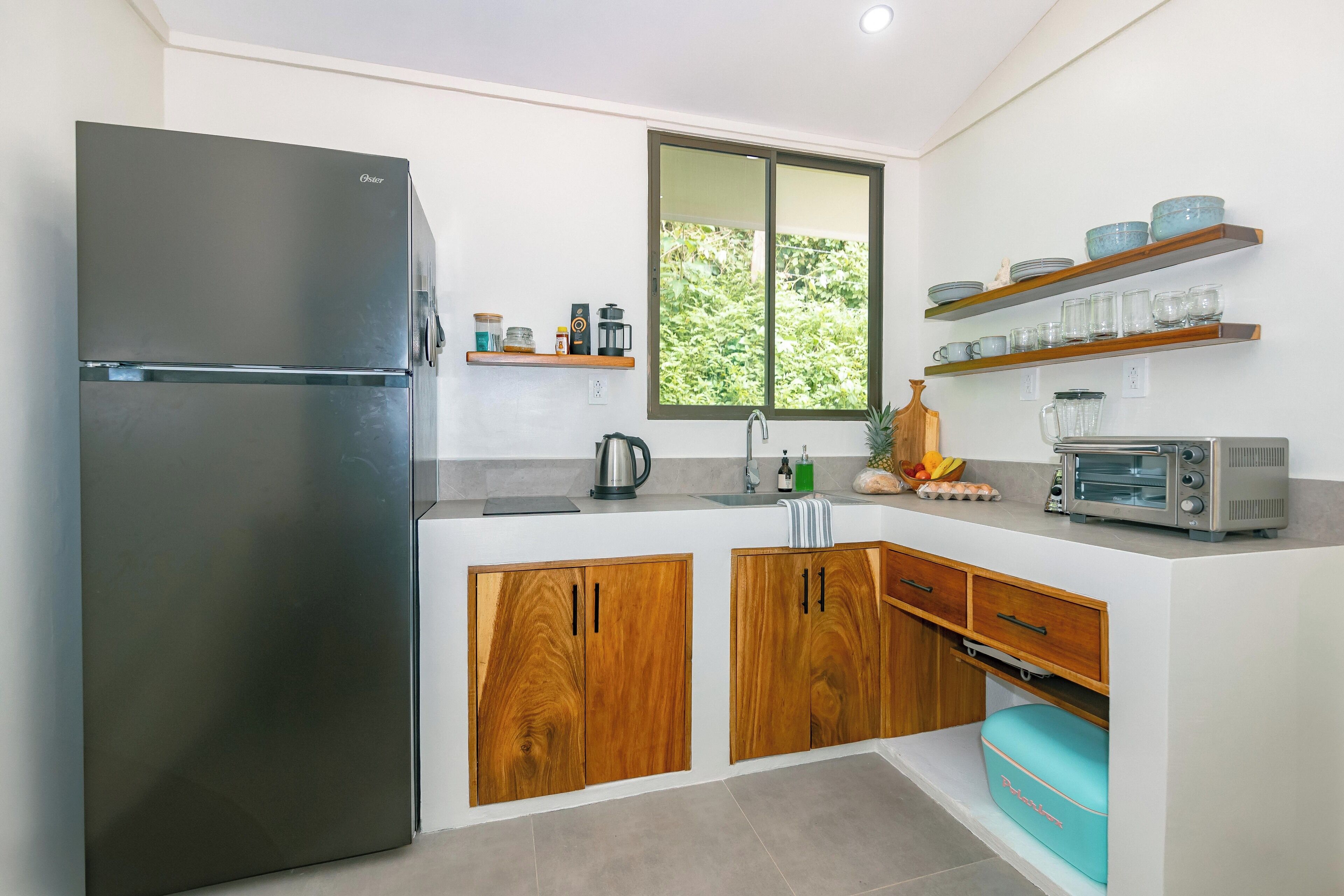 A modern kitchen with a stainless steel refrigerator, wooden cabinets, a window, and built-in shelves with dishes and a toaster.