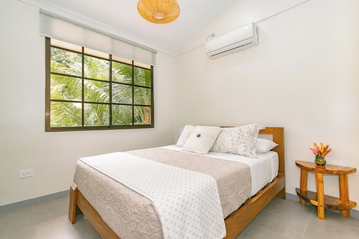 A bedroom with a large window, a bed with a wooden frame, a bedside table with a vase, and an air conditioning unit on the wall.