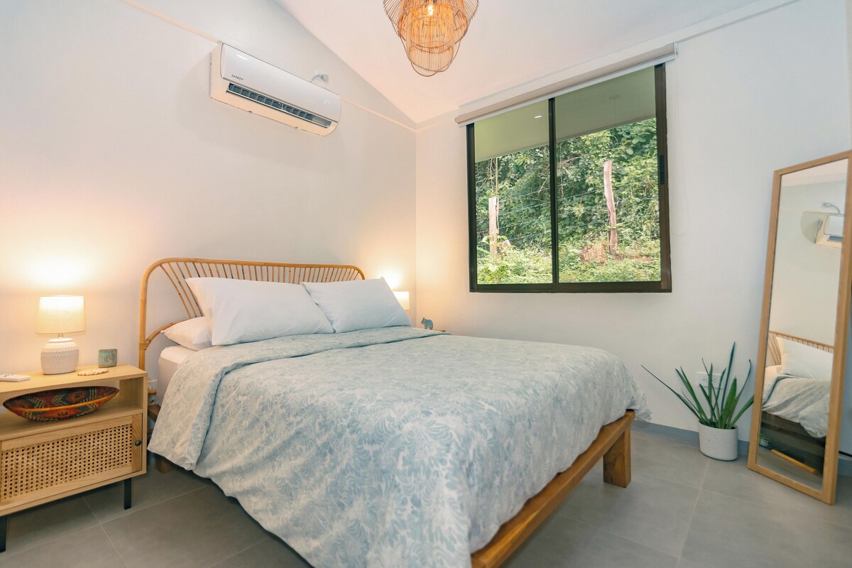 A bedroom with a bed, bedside table, lamp, air conditioning unit, window, and a potted plant.