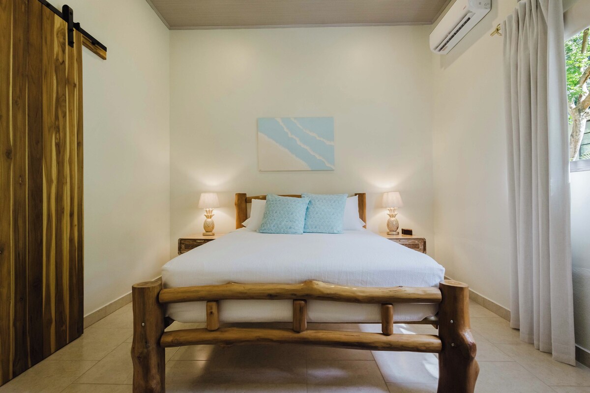 A wooden-framed bed with white bedding, two bedside lamps, a wall-mounted air conditioner, and a window with curtains.