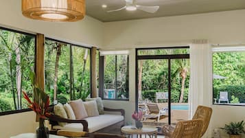 A room with a sofa, chairs, a dining table, and large windows offering a view of greenery.