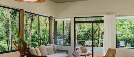 A room with a sofa, chairs, a dining table, and large windows offering a view of greenery.