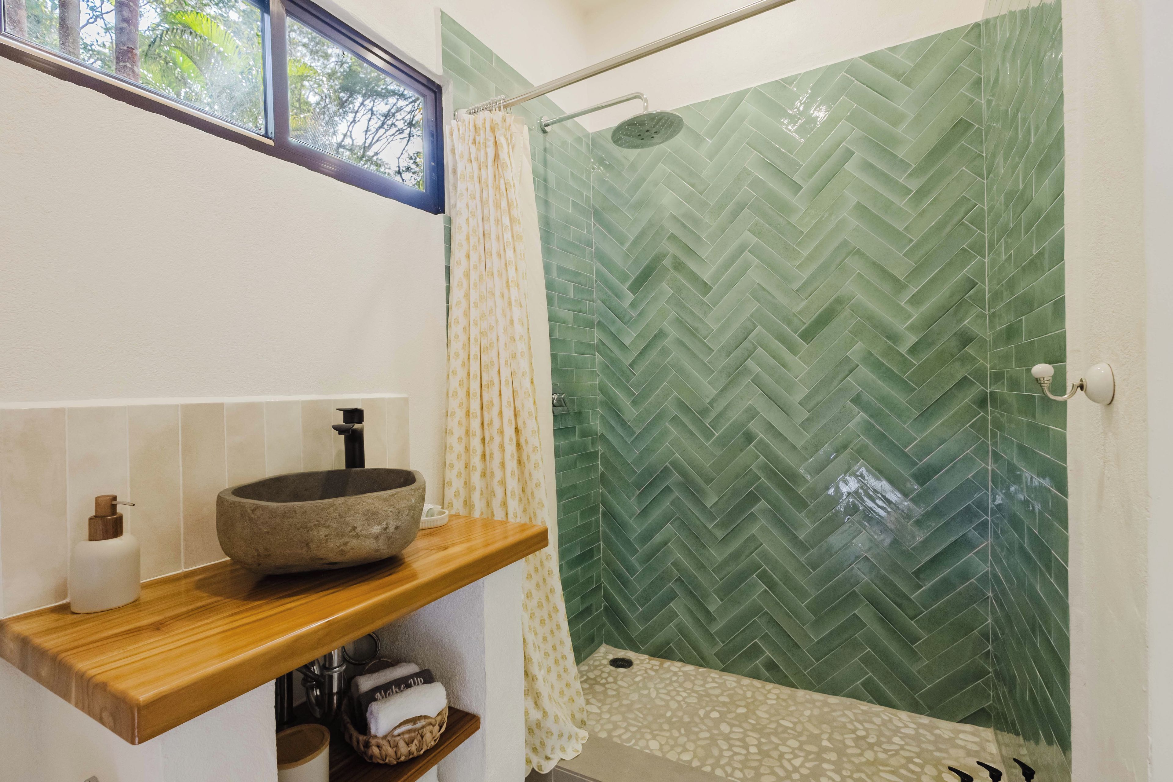 A bathroom with a stone basin, a wooden shelf, a shower with a green herringbone tile wall, and a window with a view of trees.