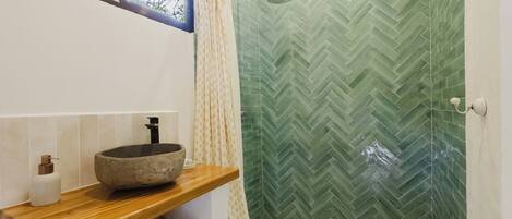 A bathroom with a stone basin, a wooden shelf, a shower with a green herringbone tile wall, and a window with a view of trees.