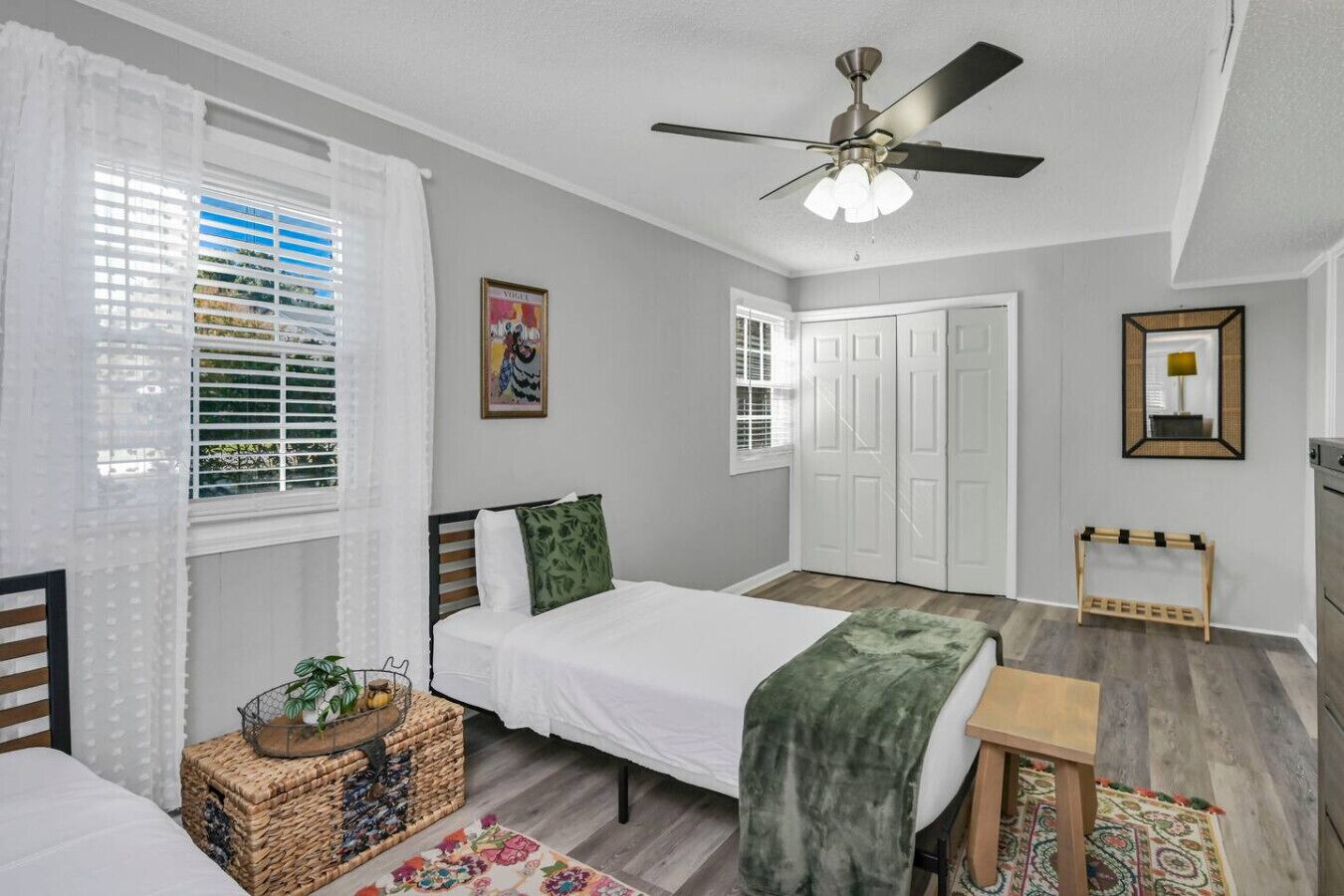 Une chambre à coucher avec un lit, un ventilateur de plafond, une fenêtre avec des stores, une porte et une petite table sur laquelle est posée une plante.