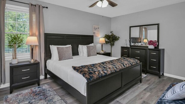 Un dormitorio moderno con cama, mesitas de noche, cómoda, ventilador de techo y un ventanal con cortinas.