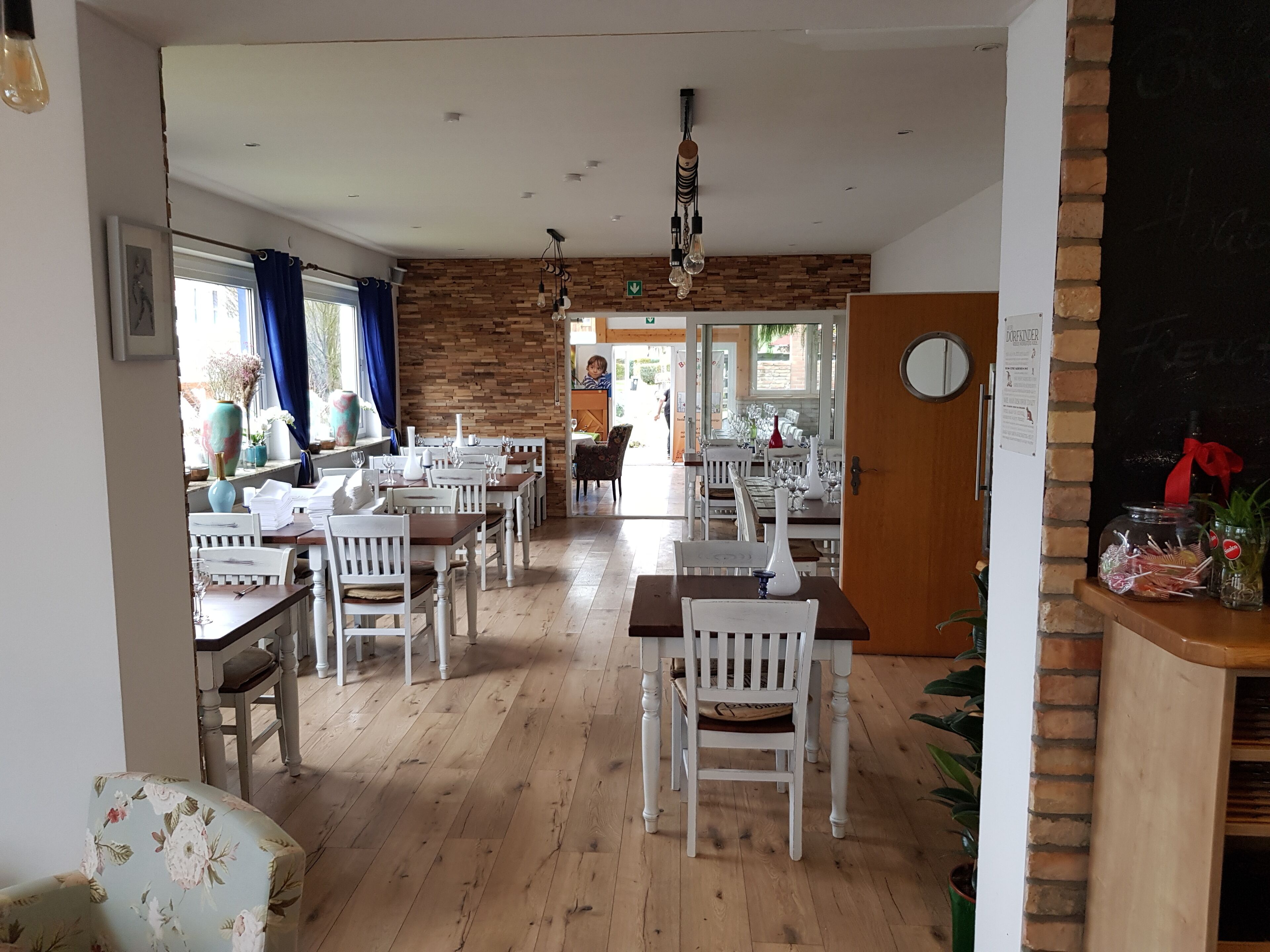 A restaurant with wooden floors, white chairs, and tables. There is a brick wall and a wooden door.