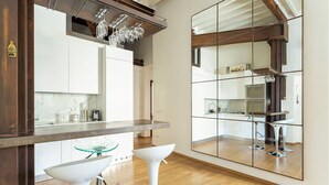Una cocina moderna con armarios blancos, una barra de vidrio, dos taburetes blancos y un espejo grande que refleja la cocina.