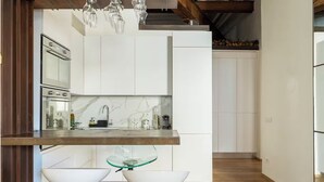 Cocina moderna con armarios blancos, salpicadero de mármol y encimera de madera. Hay dos taburetes de bar blancos y una mesa con cubierta de vidrio.
