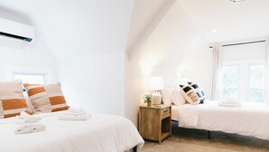 Zwei ordentlich bezogene Betten mit weißen Laken, ein Nachttisch aus Holz mit einer Lampe und ein Fenster mit Vorhängen.