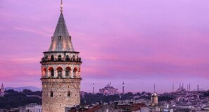 Een historisch torentje met een puntig dak en een balkon, gelegen tegen een stadsgezicht met een zonsondergang.