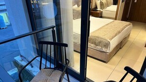 Une chambre d’hôtel avec une cloison en verre, un lit recouvert d’une courtepointe beige, une table de chevet avec une lampe et une chaise près de la fenêtre.