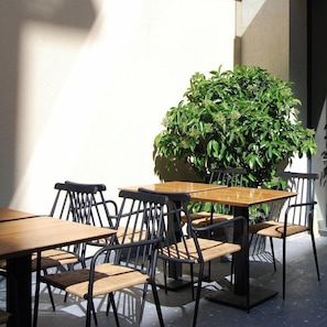 Un espacio al aire libre con mesas y sillas, una planta grande en maceta y una pared.