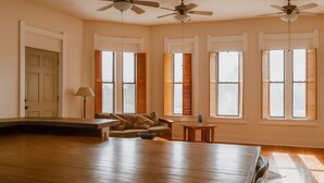 A spacious living room with hardwood flooring, a sofa, a coffee table, and a dining table. There are four windows with shutters, a door, and ceiling fans. - 15% off Spring Special | Historic Bank | Wi-Fi | Close to Purdue (Oxford)