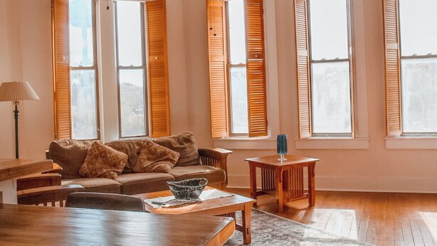 A spacious living room with a brown sofa, wooden coffee table, and a small side table. Large windows with wooden shutters allow natural light to fill the room. - 15% off Spring Special | Historic Bank | Wi-Fi | Close to Purdue (Oxford)