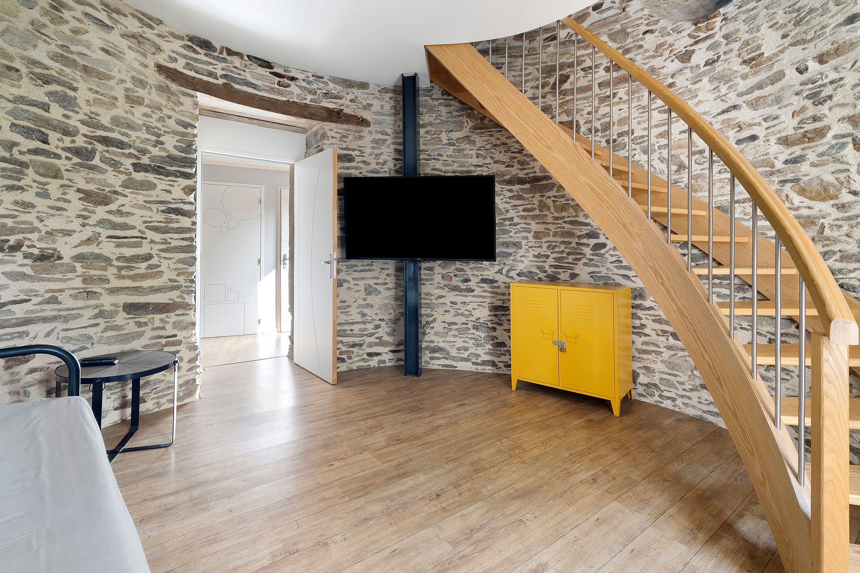 A room with a stone wall, a wooden staircase, a flat-screen TV, a yellow cabinet, and a small round table.